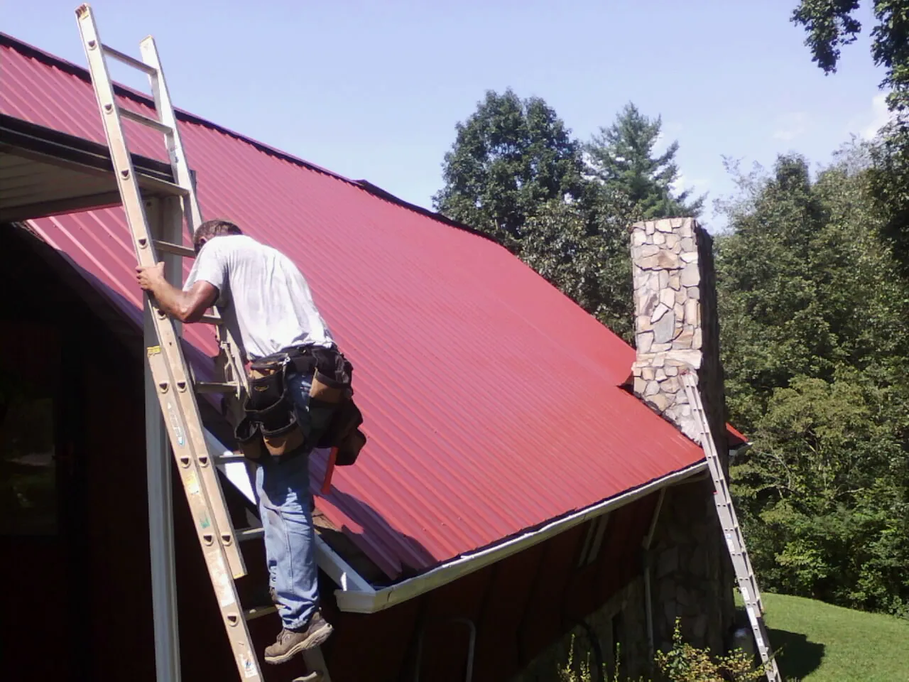 Roofer on a ladder beside a red metal roof and stone chimney — Soffit & Fascia Repair by Roofing Brothers in Montoursville